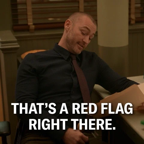 A man in a black shirt and brown tie sits at a desk reading a file. He says, 