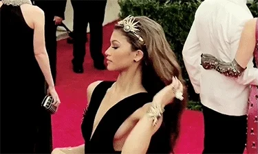Zendaya posing on a red carpet. She wears a tiara and runs her hands through her hair.
