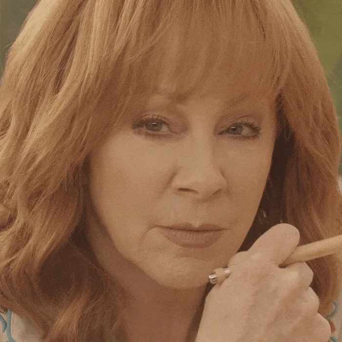 Reba McEntire squinting menacingly and suspiciously at you as she taps a pencil in her hand thoughtfully.