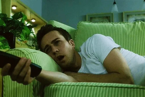 Guy lying on a green couch and holding the remote, looking zoned out and unmotivated.