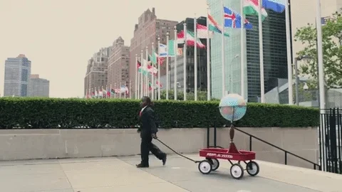 A kid in a suit carrying a globe on a wagon outside of the United Nations building in New York.