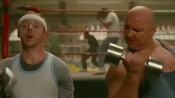 Two men lift weights together in a boxing gym.