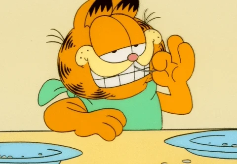 Garfield the cat sits with empty plates in front of him and moves a toothpick around his front teeth while blinking.