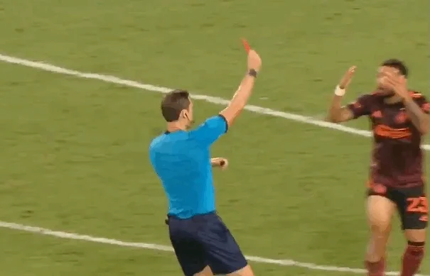 A soccer referee holds up a red card. A player reacts negatively.