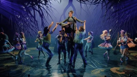 Dancers freeze while holding up one actor who sings as the camera zooms in on her