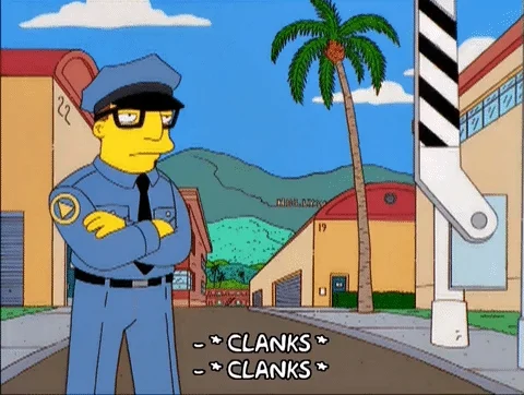 A security guard standing from The Simpsons with folded arms in front of a barrier arm gate.