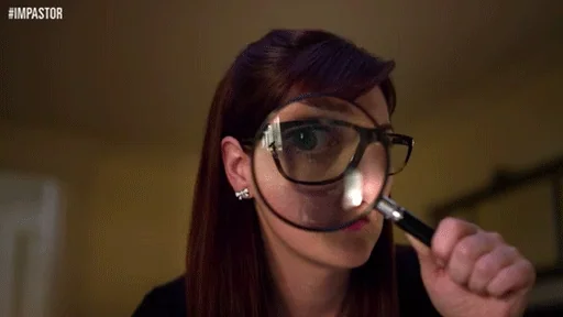 A brown haired woman with pale skin wearing black rimmed glasses looking through a magnifying glass.