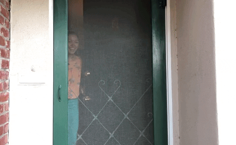 A woman opens a screen door and invites someone in.