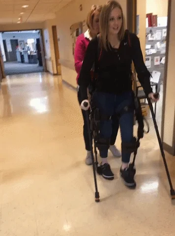 A patient walking with an exoskeleton device, assisted by a caregiver.