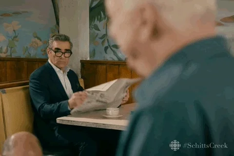 Johnny Rose from Schitt's Creek pulls a newspaper up to cover his face avoiding another character. 