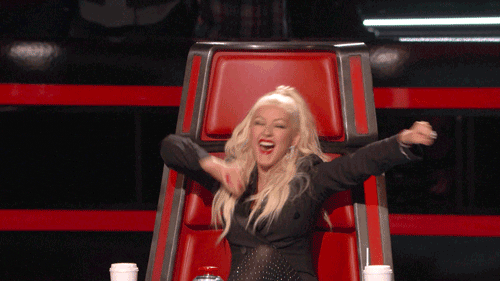 Christina Aguilera is sitting in a chair and is excitedly waving her hands in the air looking happy