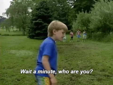 A young boy with a blue T-shirt stands in a grassy field, looking at the camera and saying, "Wait a minute, who are you?"