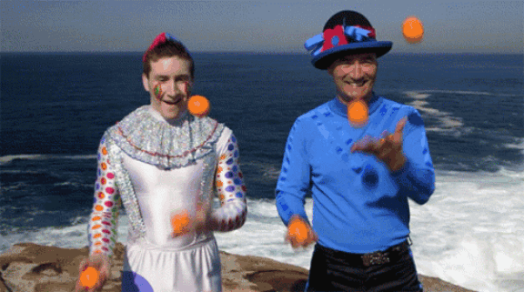 Two men juggling small balls. One dressed in a white clown suit.  One wearing a funny black hat with blue-red decorations.