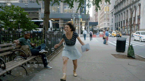A woman happily skipping down a sidewalk as people sitting on a bench with bubble guns shoot out bubbles toward her. 