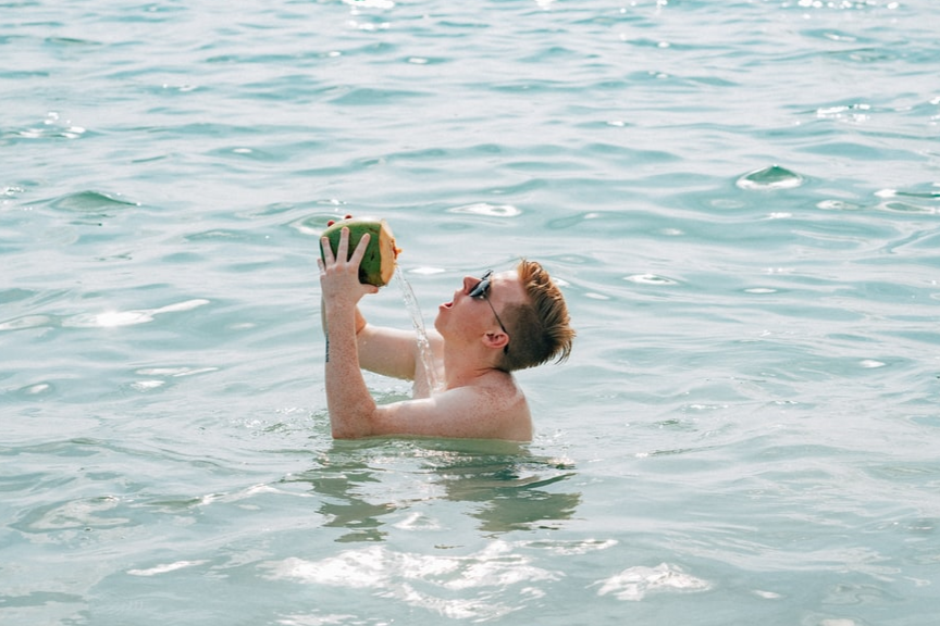 Image of man chest-deep in seawater failing to drink from coconut.