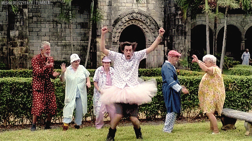 Jim Carey dancing in a tutu. Elderly people dance behind him on a lawn.