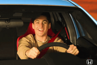 A young man smiles joyfully with both his hands on the steering wheel.