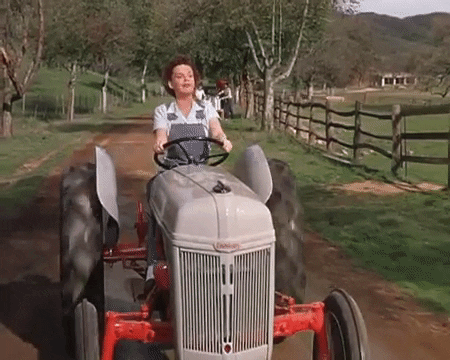 A woman wearing overalls drives a tractor and sings