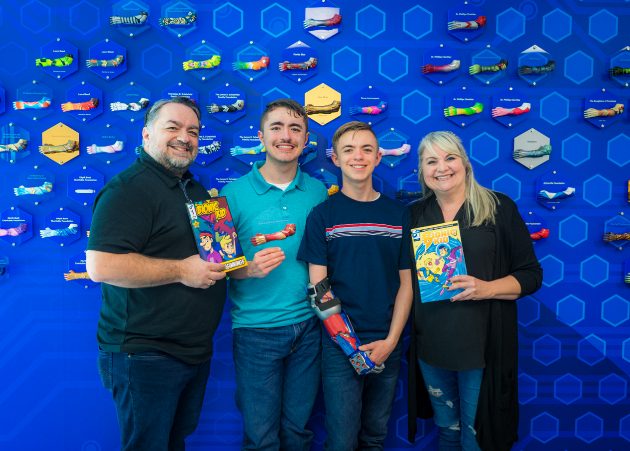 An image of a bionic child with his family holding The Bionic Kid comic books and a donor plaque. They are in front of a donor recognition wall with mounted mini arms.