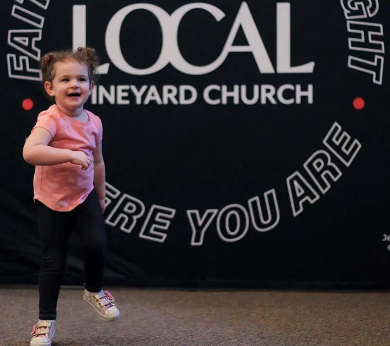 a smiling child running toward a camera with 'the local vineyard church' sign in the background