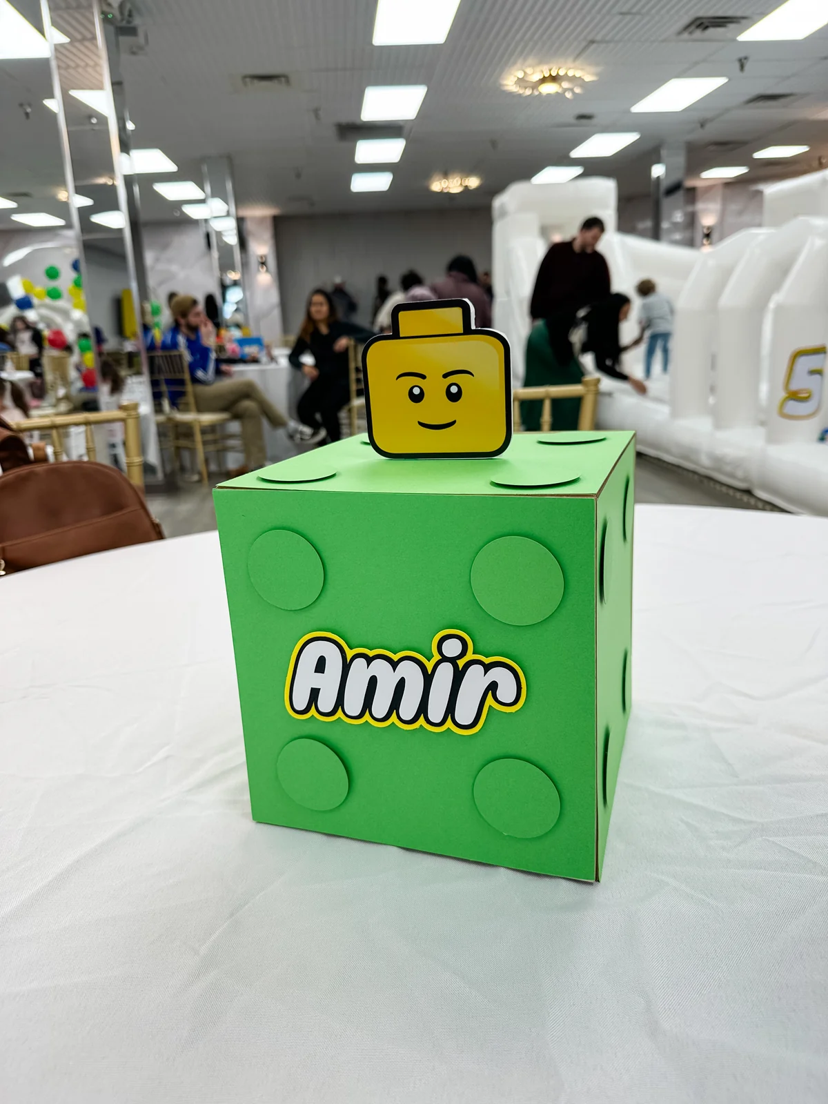 Green Lego-themed party centerpiece box with bounce house and guests in background