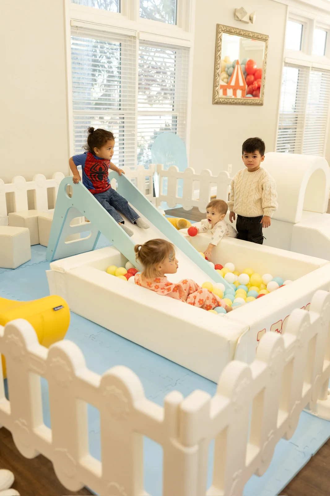 Children sliding into a colorful ball pit with soft play equipment in a bright indoor space