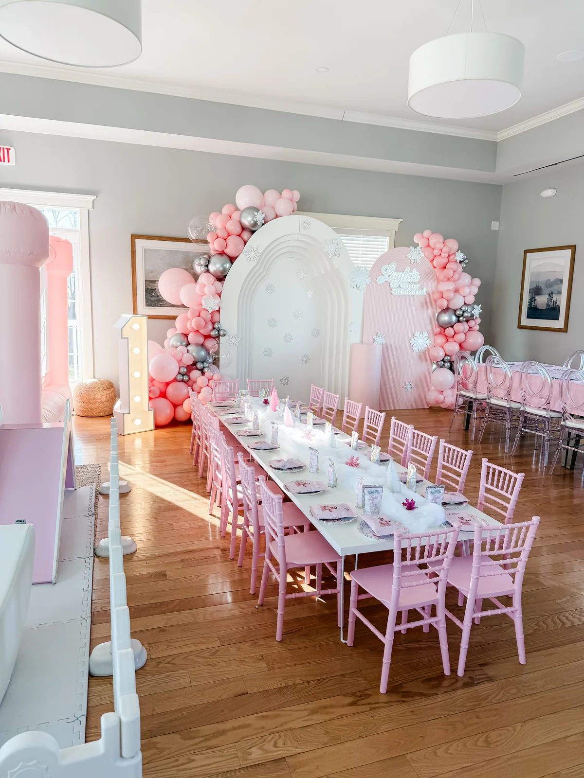 Pink birthday party room with long table, balloon arch, white arch backdrop, and marquee number