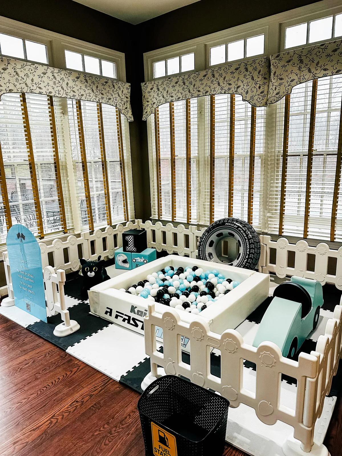 Racing-themed soft play with black and teal ball pit, toy tire props, and play car in sunroom