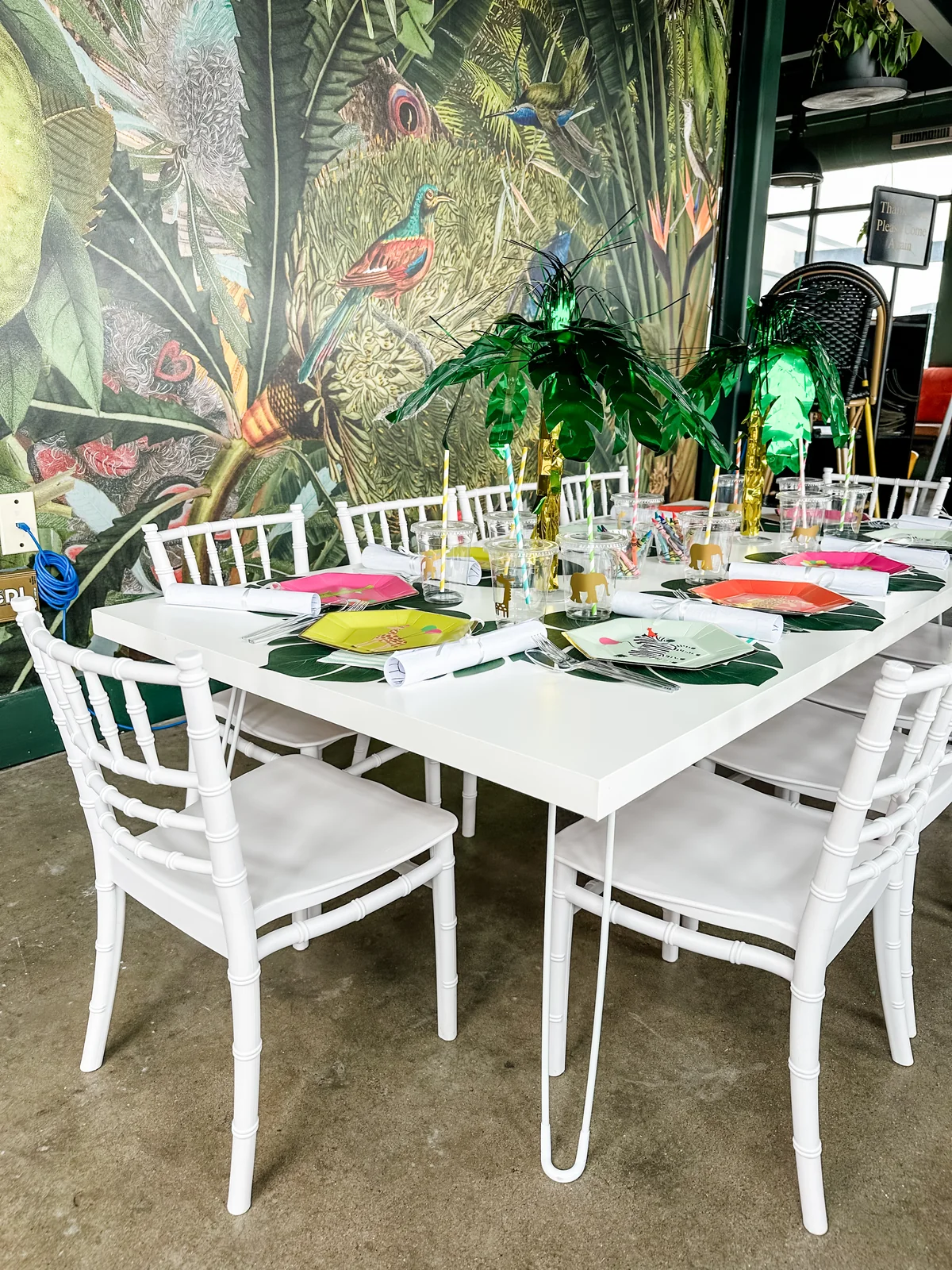 Tropical-themed tablescape with white chairs and palm centerpiece against jungle mural
