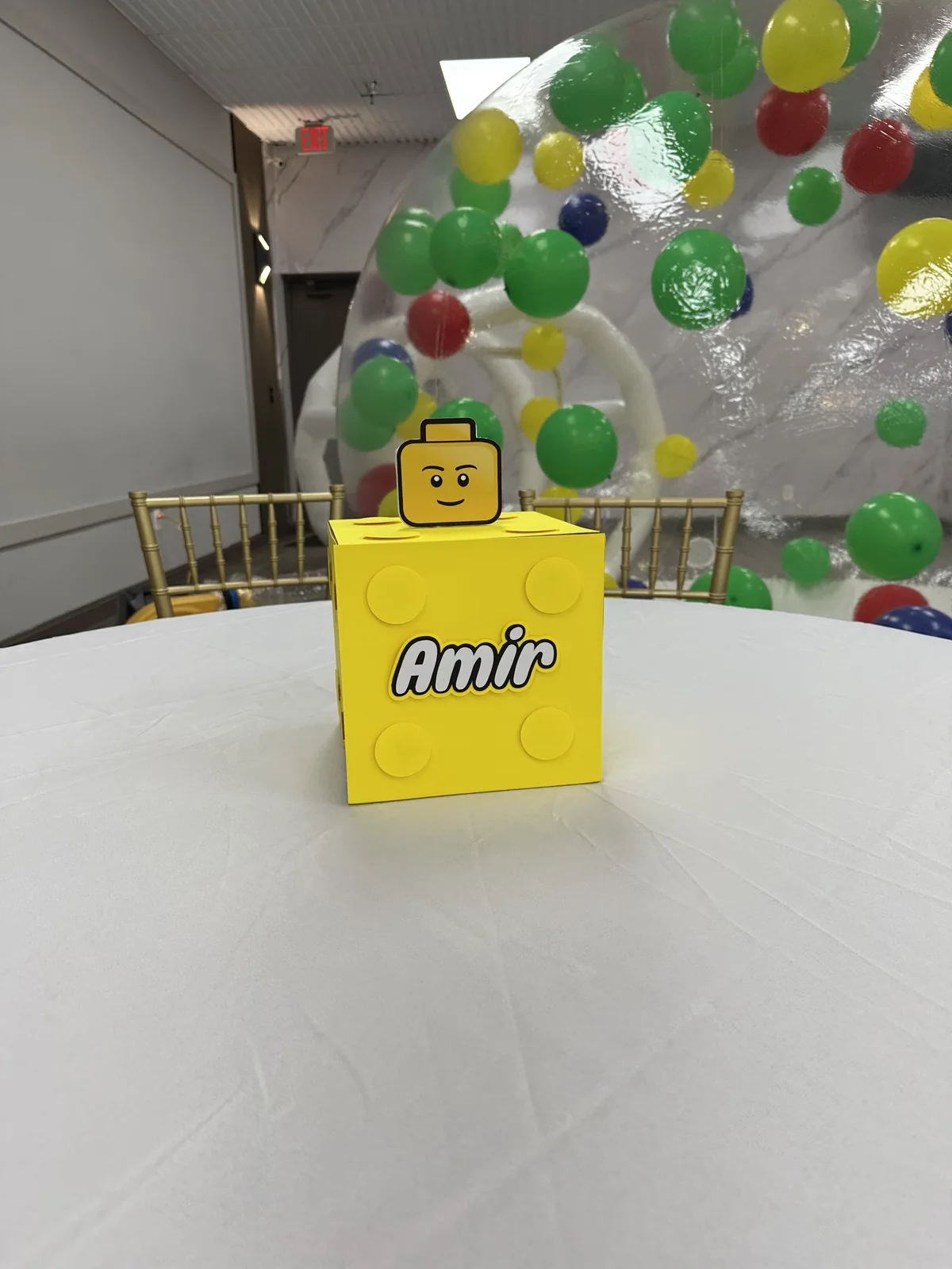 Lego-themed custom favor box on party table with colorful balloon arch in background