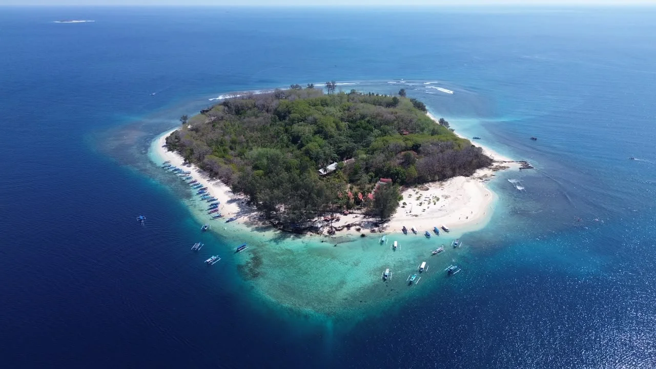 Pemandangan udara Gili Nanggu dengan pantai pasir putih, hutan rindang, dan perairan toska yang dikelilingi perahu.