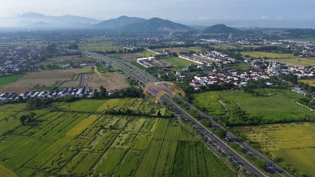 Pemandangan udara Kota Mataram dengan jalan raya, gapura warna-warni, permukiman, sawah, dan perbukitan di kejauhan.