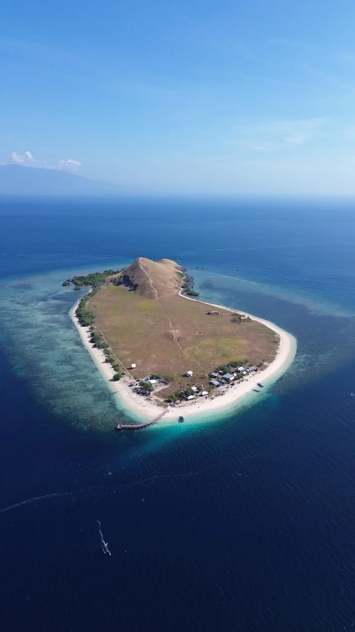 Pemandangan udara Pulau Kenawa dengan bukit savana, pantai pasir putih, dan dermaga kayu