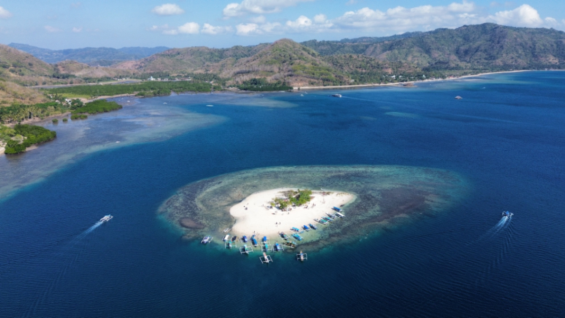 Pemandangan udara Gili Kedis dengan hamparan pasir putih, terumbu karang, perahu tradisional, dan perbukitan Sekotong.