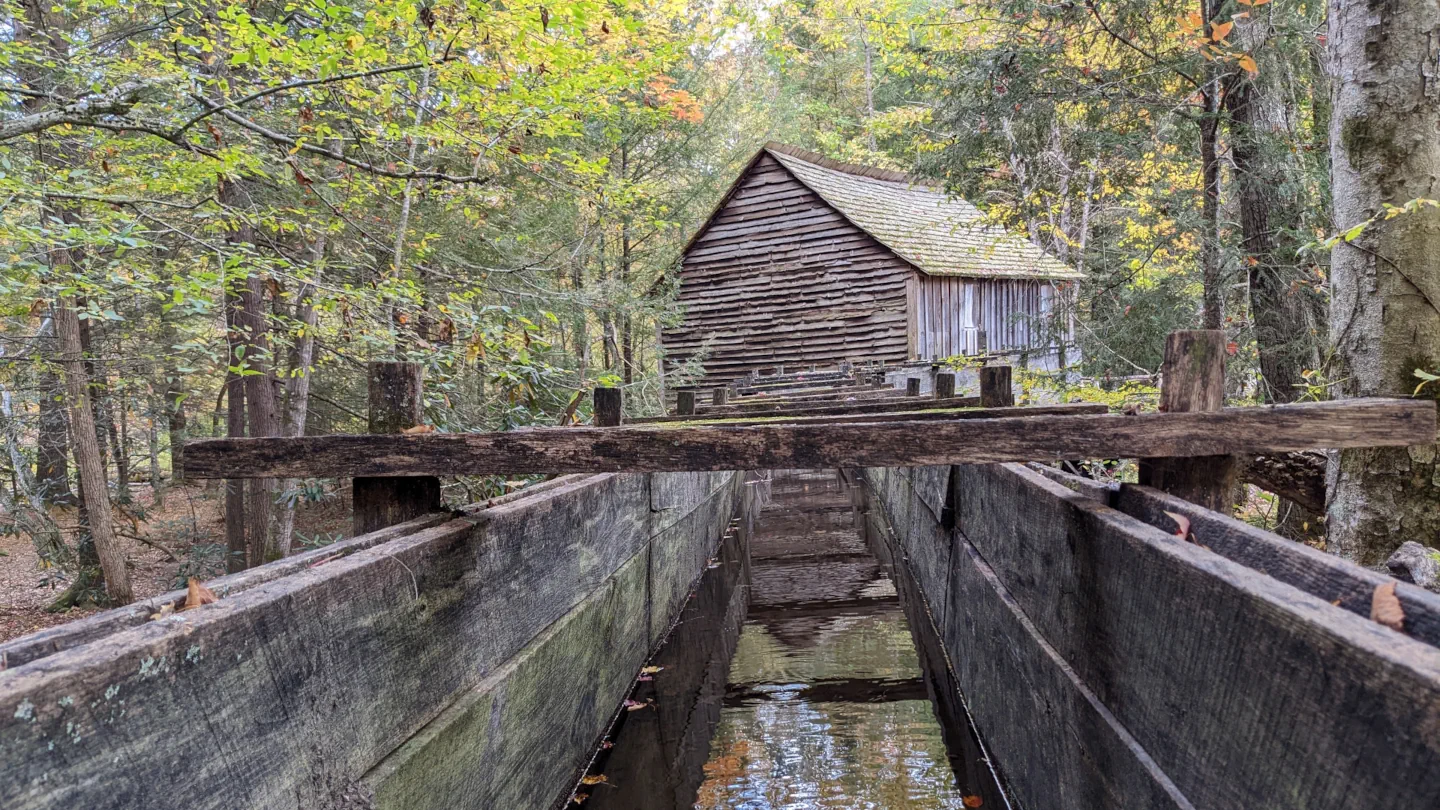 Photo of an old wooden barn in the woods towards the end of summer, just as the leaves are starting to change. The perspective is peering down a long  wooden trough or canal filled with water. The trees, sky, and barn reflect against the still water.