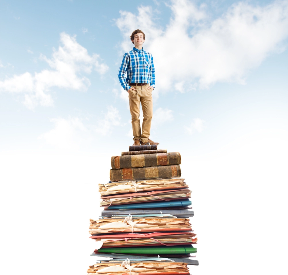 A young man in a blue and white plaid shirt and khakis, standing on top of a giant pile of books, folders, and laptops in front of a blue cloudy sky.
