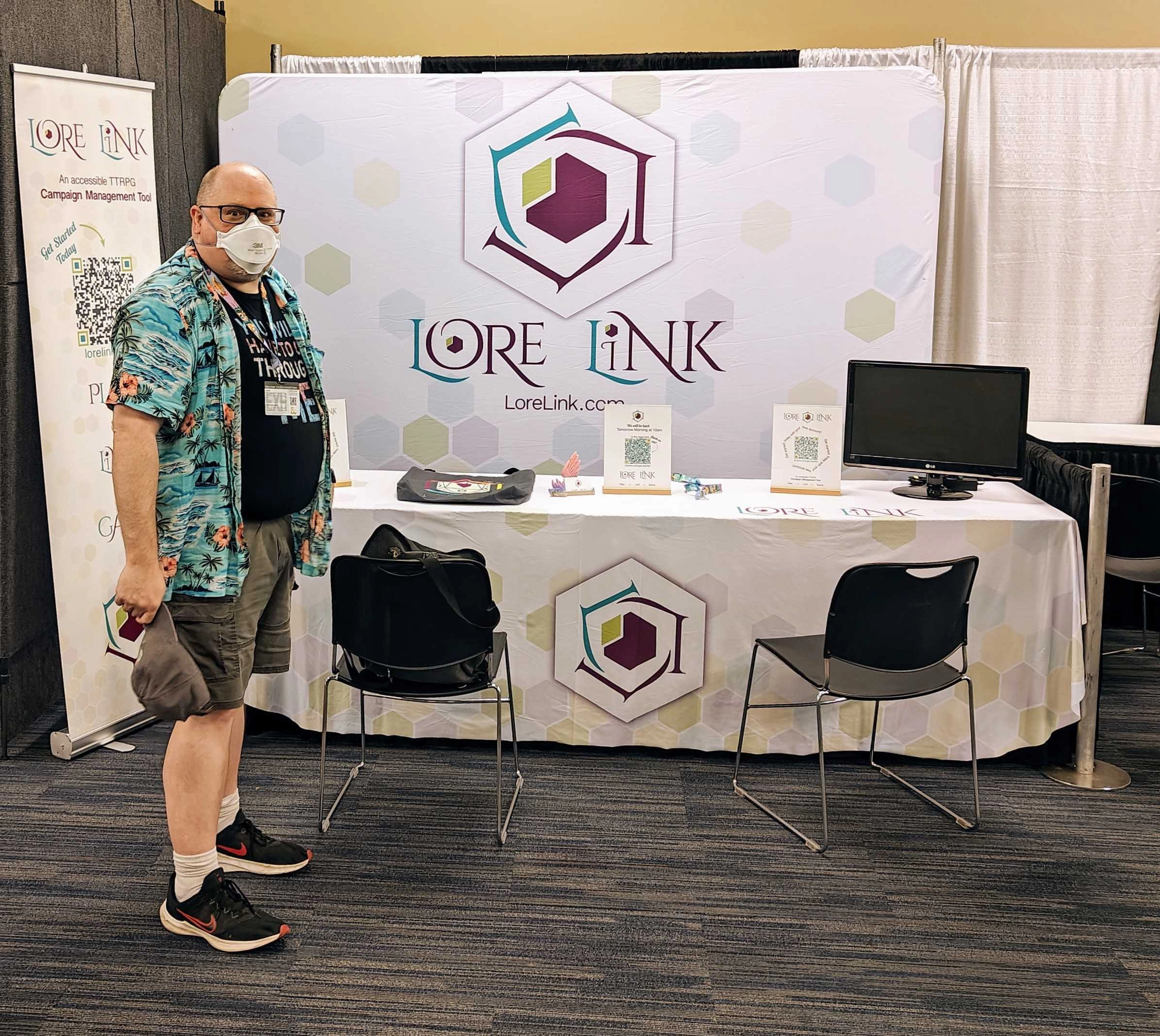 A bald man wearing a mask, looking at the camera while standing in front of the Lore Link booth at Gen Con.