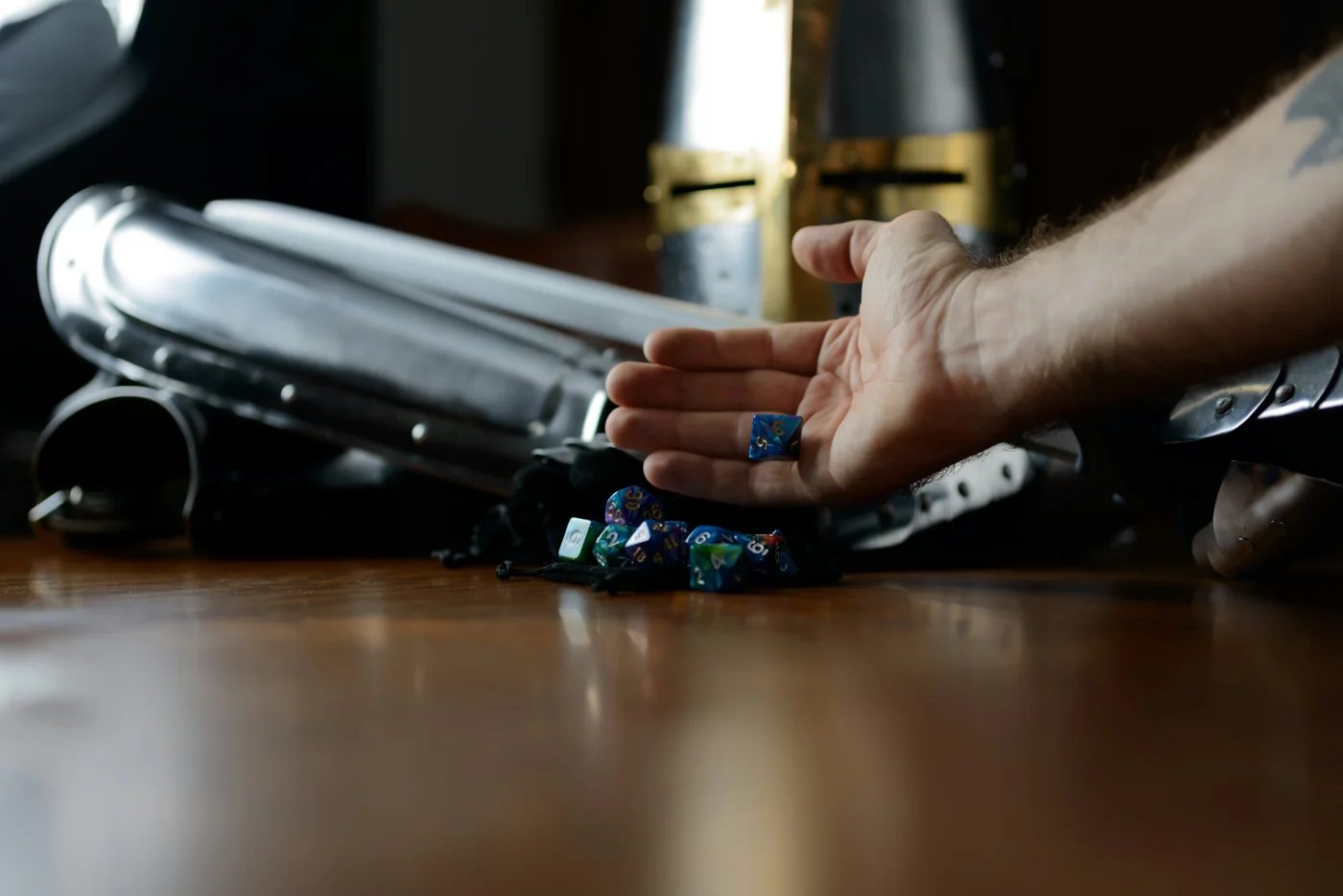 A hand is reaching across a wooden table to toss dice. On the table and behind the hand is a suit of silver-colored armor. A great helm with gold colored accents features prominently and in the display.