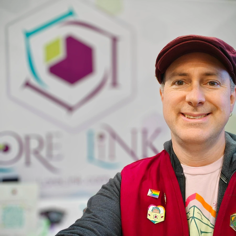 Michael in front of the Lore Link booth at Origins 2024, wearing a deep red vest and hat, and pink Lore Link T-shirt. Pinned on the vest are a Progress Pride flag and the heart-shaped Lore Link Gamemaster badge.