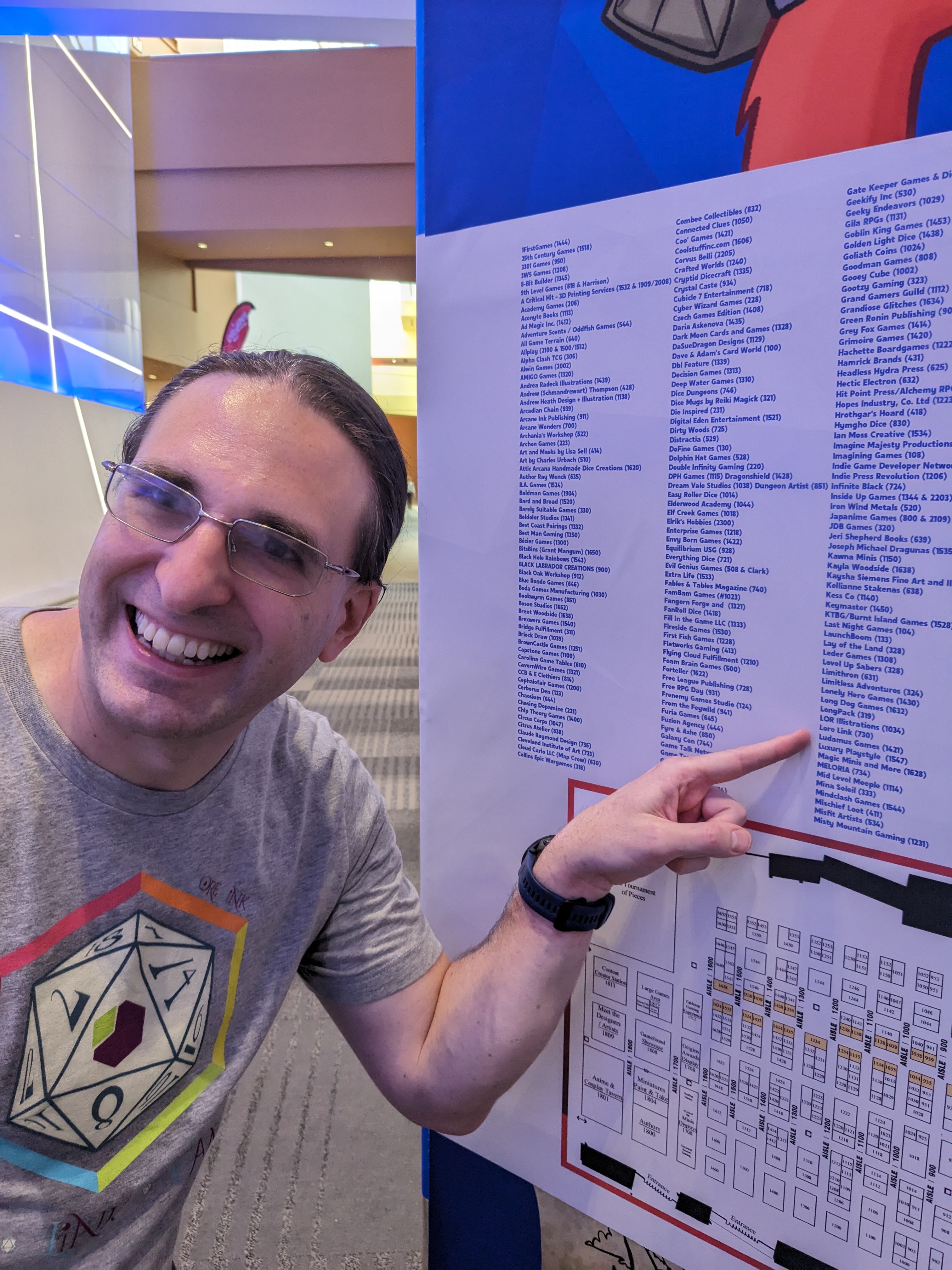 Mark, the mind behind Lore Link, grinning and pointing to Lore Link on the long list of vendors at Origins 2023.