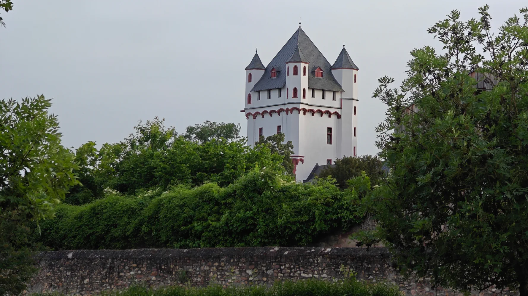 A white tower with round  towers in each corner rises above a stone wall trimmed with shrubs and ivy. The tower features red accents and slate grey peaked roof. Beside the tower is a wooden building largely obstructed by the trees.