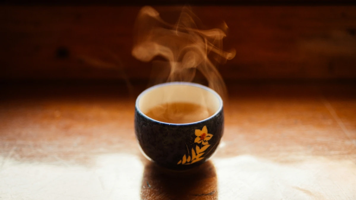 Steam rises from a simple dark teacup decorated with a white flower and resting on a wooden table.