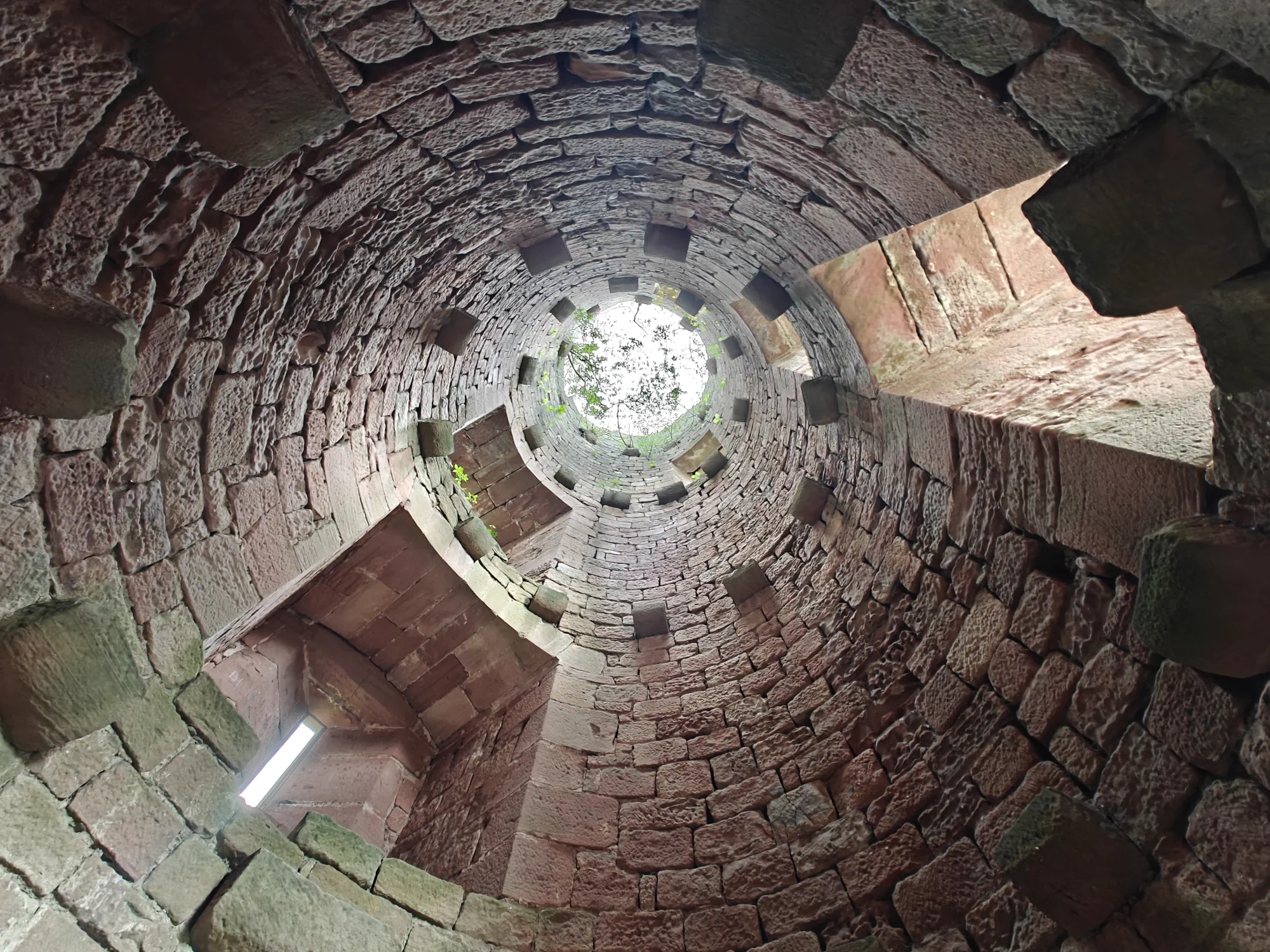 A photo of the sky from the basement of an ancient round tower of the Landsberg Castle. Arrows slits are visible in one window with stonework spiraling around the viewer to dizzying height, with tree branches visible through the opening at the sky.