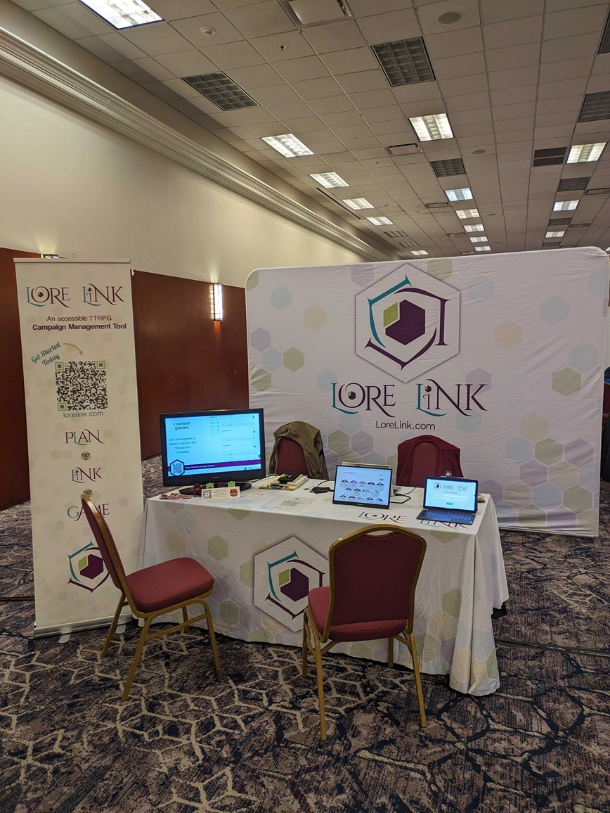 The Lore Link booth at Who's Yer Con. There is a backdrop with a large Lore Link logo on it, with a pair of chairs behind a table with three screens for demos. In front of the table, which is covered in a tablecloth with the Lore Link logo on it as well, are a pair of chairs for people to watch demos, and a banner with a QR code.