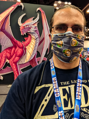 Gencon A man with glasses and a mask over his mouth and nose, pictured in front of a cutout of a red dragon