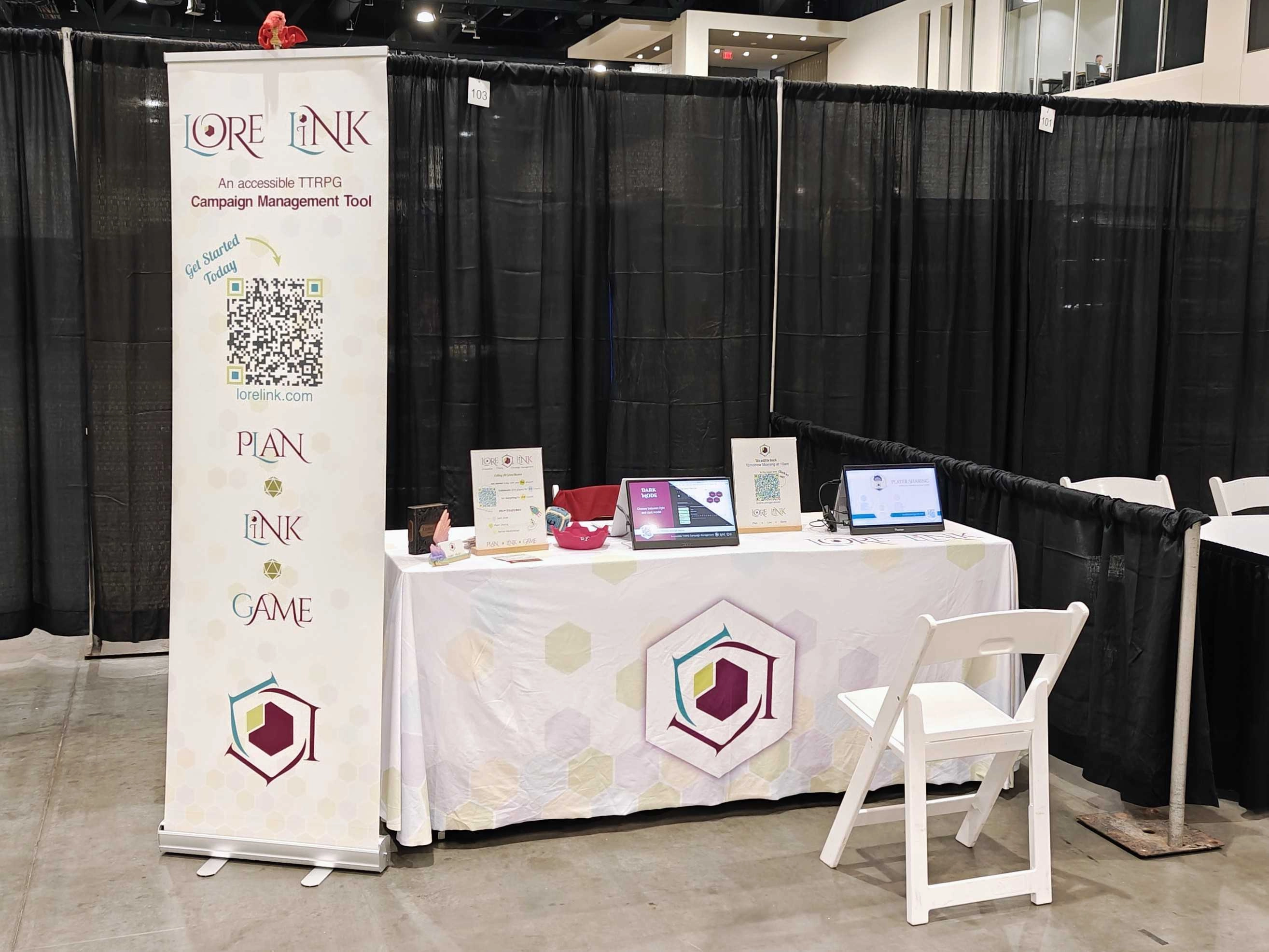 A picture of the simple booth set up for Playthrough GC. We have a banner at the end of our table, and the table has the Lore Link tablecloth covering it, and it has a pair of portable monitors for demonstration, signage with QR codes, and small giveaway items.