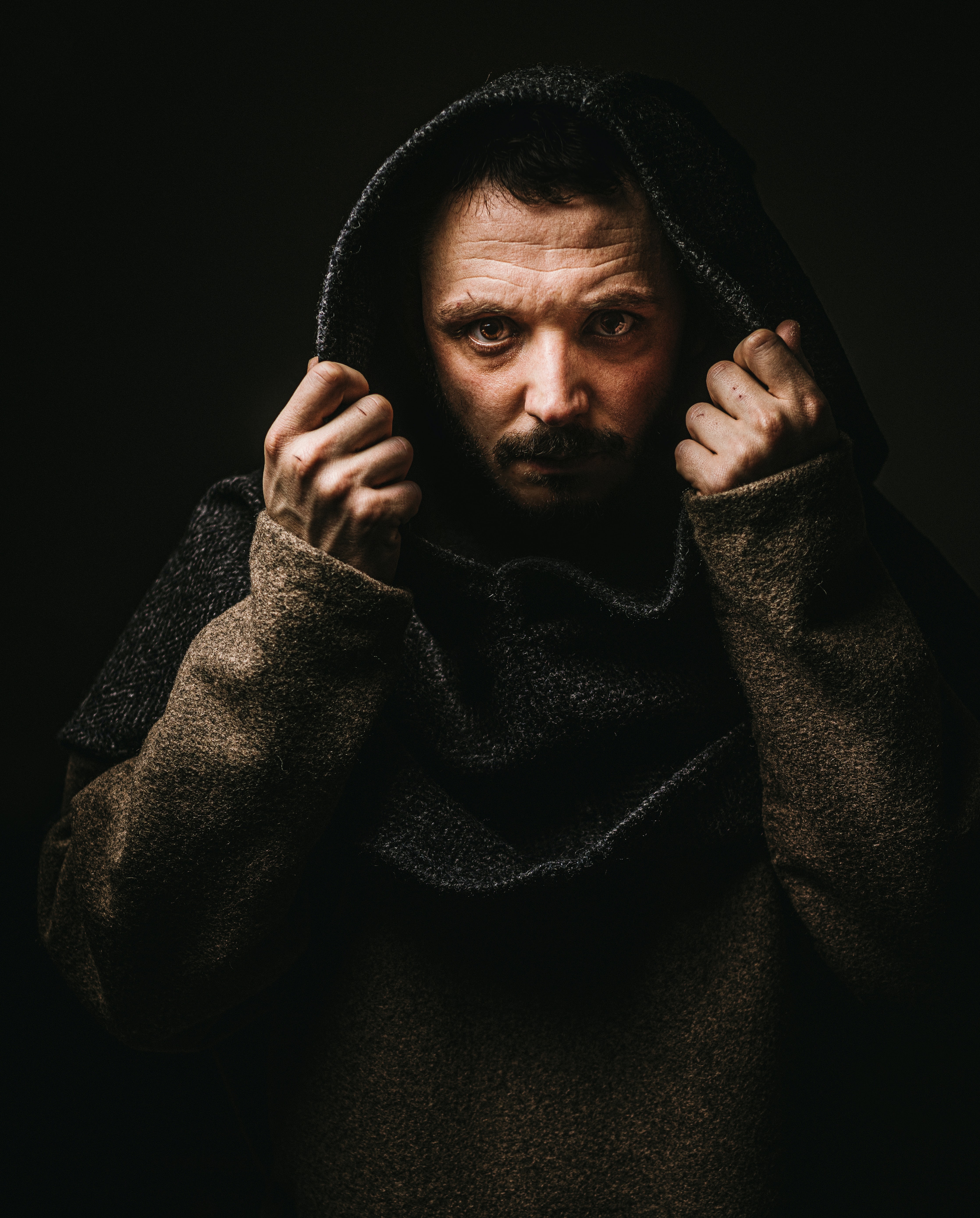 A photo of a man, looking at the camera, pulls a hood over his head against a black background