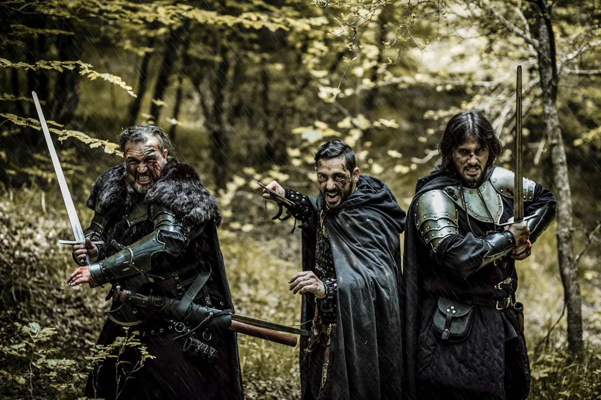 A party of three adventurers in the woods, each with serious expressions and swords drawn as if to charge into combat. The are wearing various armor, cloaks, and furs, along with pouches and other accessories.