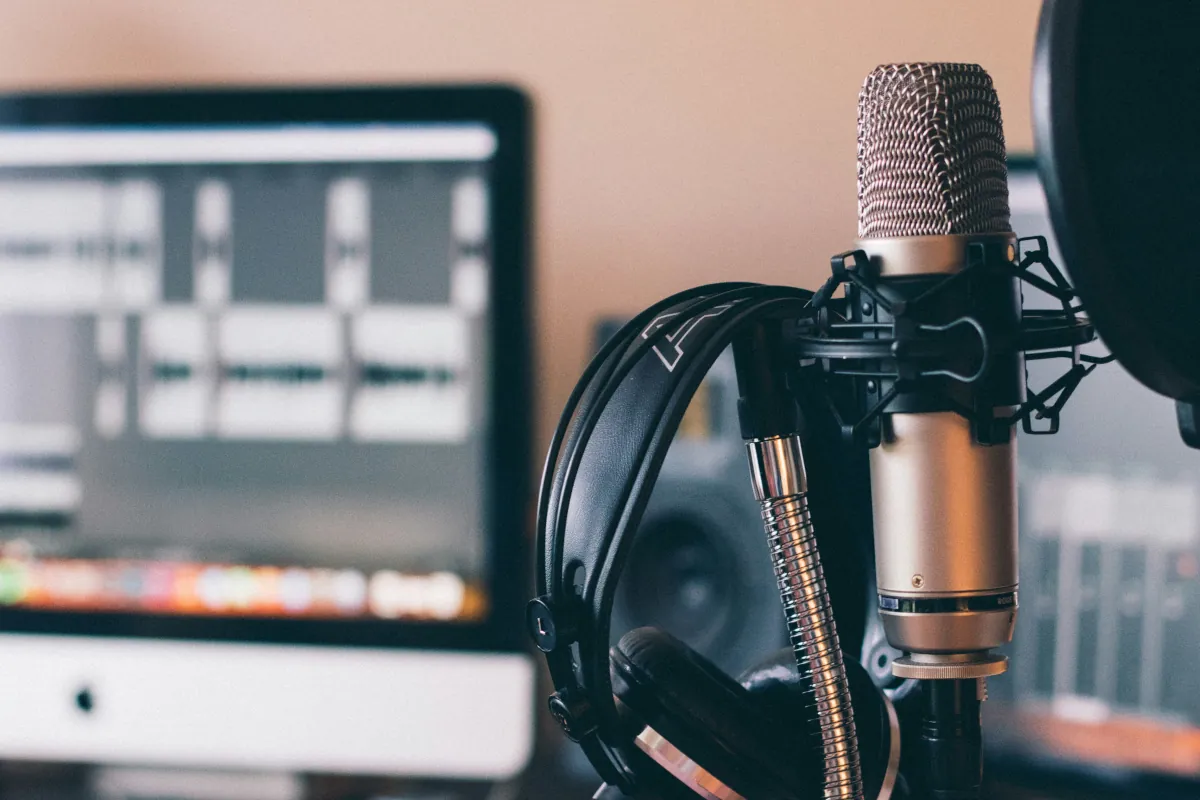 A podcasting and recording setup is shown with a studio microphone and pop filter in the foreground, a headset is hanging from a stand, and in the background a large screen is showing audio files in the process of editing.