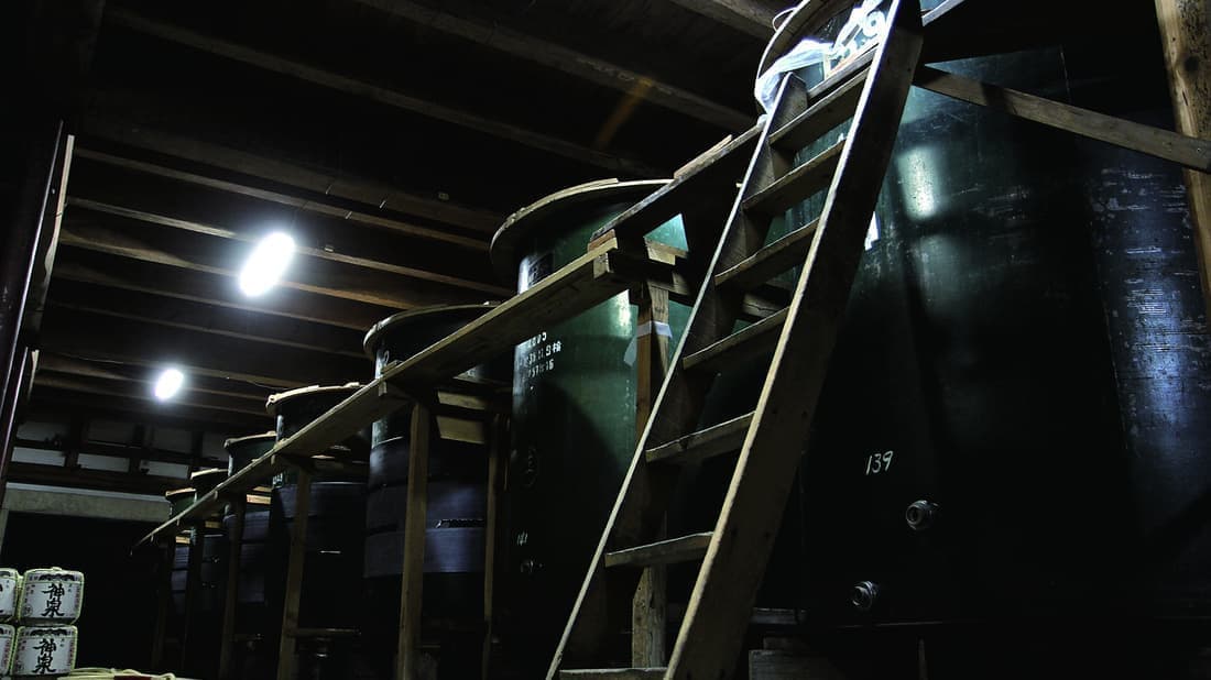 A wooden ladder leads up to large dark fermentation tanks stacked in the brewery's storage building.